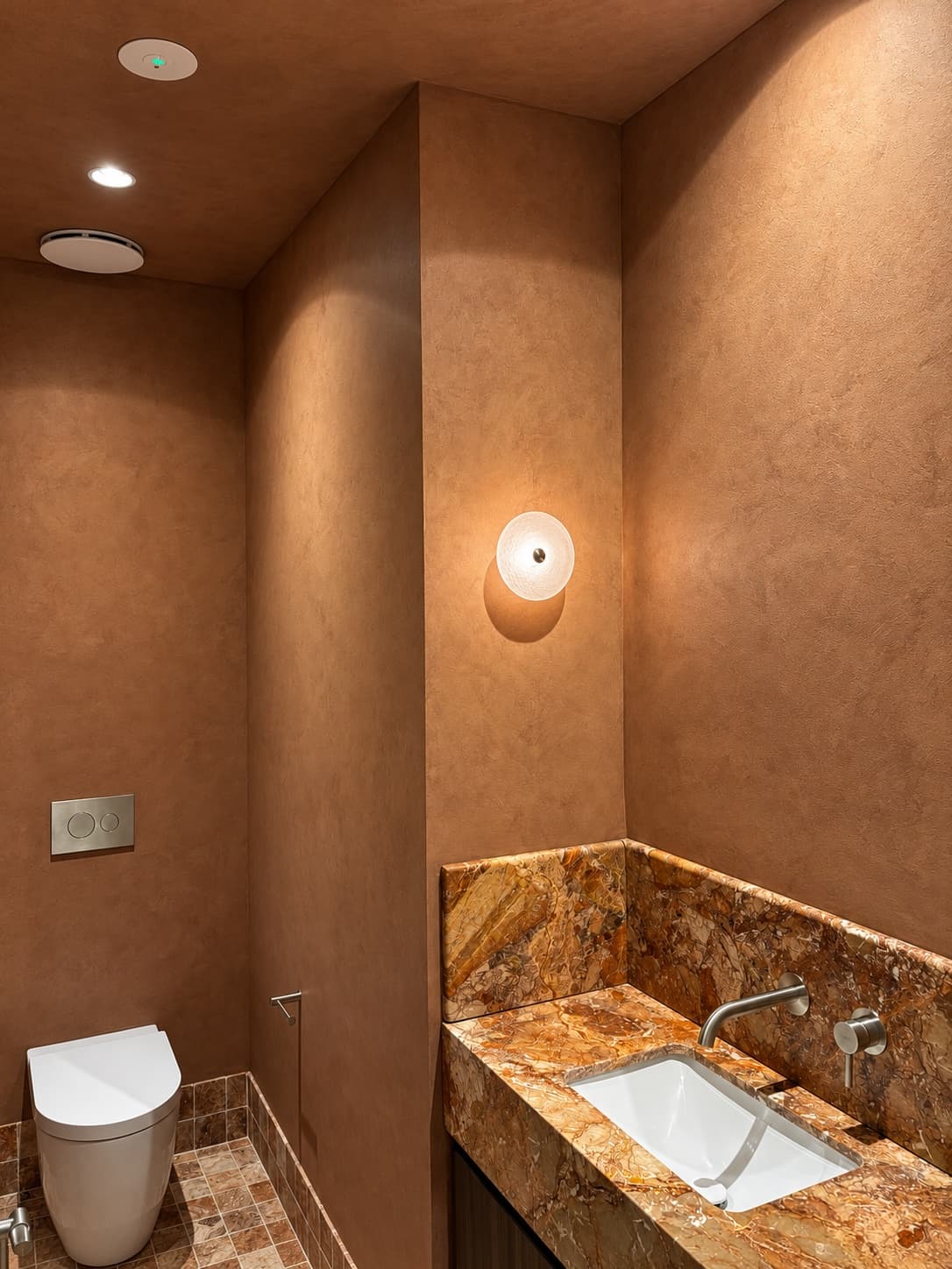 Terracotta French wash across the walls and ceiling of a washroom, lit by a glass wall sconce.